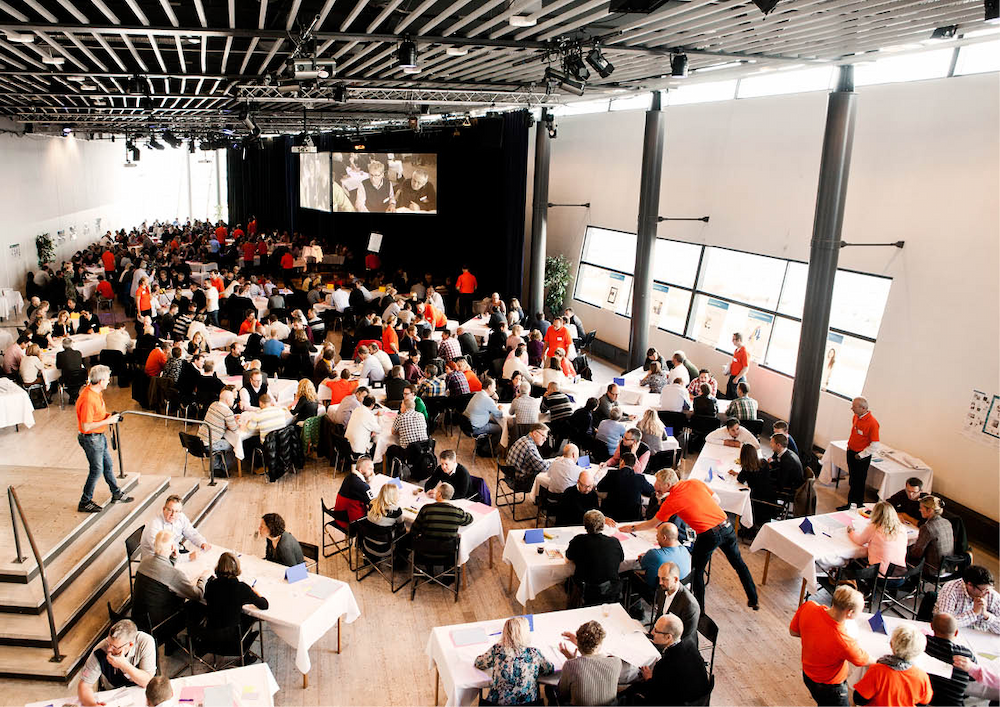 A large group of people are seated at tables in a brightly lit conference hall, with some individuals standing and interacting.
